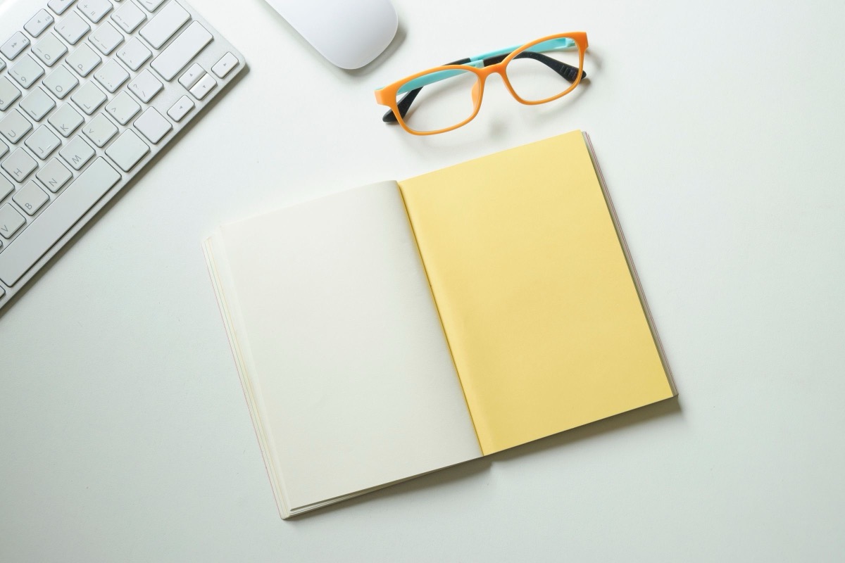 A notebook, glasses and a keyboard on a white desk